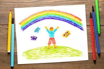 The boy rejoices in the rainbow. Children's drawing on white paper. Wooden table with markers