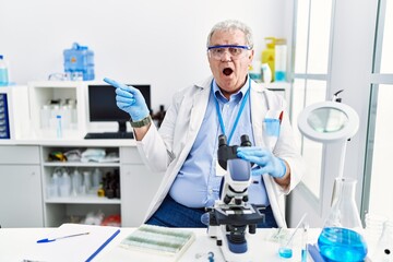 Senior caucasian man working at scientist laboratory surprised pointing with finger to the side, open mouth amazed expression.