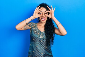 Young woman wearing bindi and traditional kurta dress doing ok gesture like binoculars sticking tongue out, eyes looking through fingers. crazy expression.