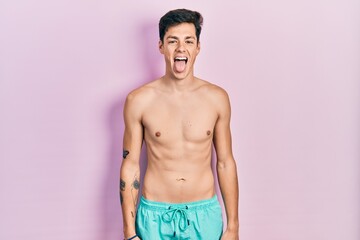 Young hispanic man wearing swimwear shirtless sticking tongue out happy with funny expression. emotion concept.