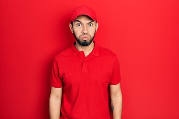 Hispanic man with beard wearing delivery uniform and cap puffing cheeks with funny face. mouth...