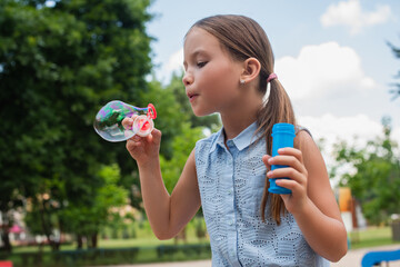 girl in sleeveless blouse blowing soap bubble in park.