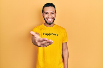 Hispanic man with beard wearing t shirt with happiness word message smiling friendly offering handshake as greeting and welcoming. successful business.