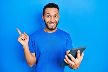 Hispanic man with beard using touchpad device smiling happy pointing with hand and finger to the side