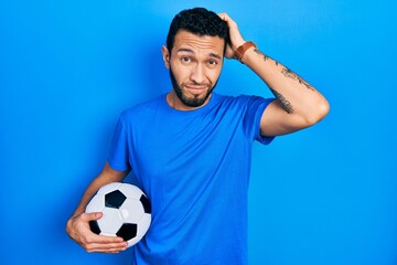 Hispanic man with beard holding soccer ball confuse and wondering about question. uncertain with...