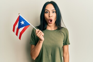 Young hispanic girl holding puerto rico flag scared and amazed with open mouth for surprise,...