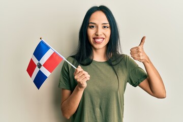 Young hispanic girl holding dominican republic flag smiling happy and positive, thumb up doing...