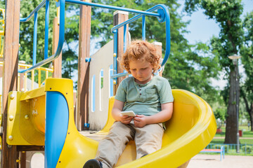 redhead boy sitting on slide in amusement park and playing on smartphone.