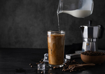 Milk from a dark jug is poured into a glass of cold coffee
