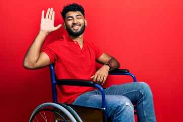 Arab man with beard sitting on wheelchair waiving saying hello happy and smiling, friendly welcome gesture