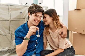 Young caucasian couple smiling happy sitting on the floor holding key of new home.
