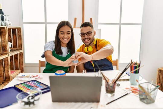 Young hispanic couple using laptop sitting on the table at art studio smiling in love showing heart symbol and shape with hands. romantic concept.