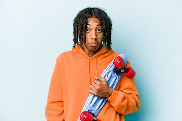 Young African American skater man isolated on blue background shrugs shoulders and open eyes...