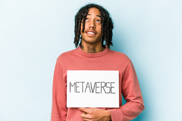 Young African American man holding metaverse placard isolated on blue background dreaming of achieving goals and purposes