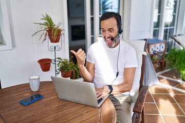 Middle age man wearing call center agent headset working from home pointing thumb up to the side smiling happy with open mouth