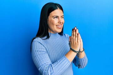 Beautiful brunette woman praying holding catholic rosary winking looking at the camera with sexy...