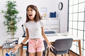 Young hispanic girl standing at pediatrician clinic angry and mad screaming frustrated and furious,...