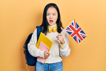 Young chinese girl exchange student holding uk flag in shock face, looking skeptical and sarcastic,...
