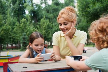 happy woman smiling near kids using smartphones in park.