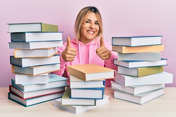 Young caucasian woman sitting on the table with books approving doing positive gesture with hand, thumbs up smiling and happy for success. winner gesture.
