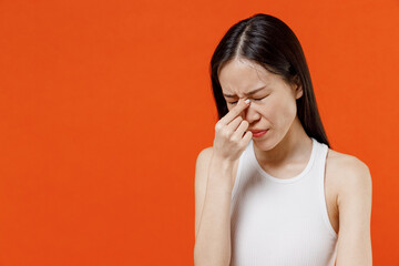 Fototapeta premium Frustrated tired exhausted unhappy preoccupied young woman of Asian ethnicity 20s years old in white tank top keep eyes closed rub put hand on nose isolated on plain orange background studio portrait.