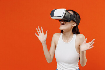 Shocked surprised fun cheerful blithesome stupefied young woman of Asian ethnicity 20s years old in white tank top watching in vr headset pc gadget isolated on plain orange background studio portrait.