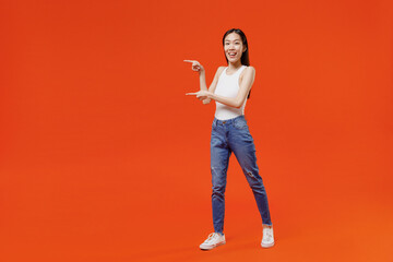 Full size body length side view young woman of Asian ethnicity 20s in white tank top pointing forefingers away on workspace area copy space mock up isolated on plain orange background studio portrait.