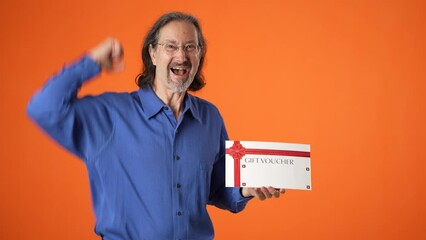 Happy mature man 50s 60s hold gift certificate voucher tracking points winner gesture looking camera isolated on orange color background studio portrait.