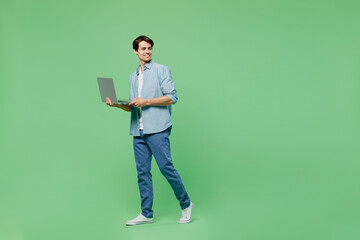 Full size body length side view smiling young brunet man 20s years old wears blue shirt hold use work on laptop pc computer looking aside step walk isolated on plain green background studio portrait. - obrazy, fototapety, plakaty