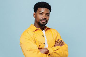 Young confident happy attractive man of African American ethnicity in yellow shirt hold hands crossed folded isolated on plain pastel light blue background studio portrait. People lifestyle concept