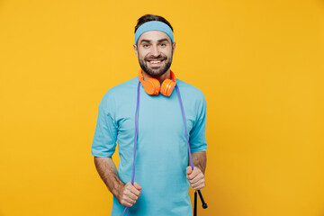 Young fun fitness trainer instructor sporty man sportsman in headband blue t-shirt spend weekend in home gym hold skipping rope isolated on plain yellow background. Workout sport motivation concept.