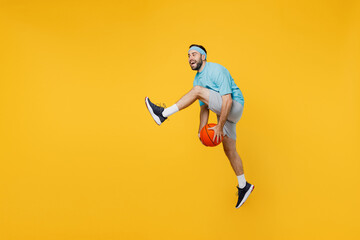 Fototapeta na wymiar Full size caucasian young fitness trainer instructor sporty man sportsman in headband blue t-shirt hold ball play basketball game jump high isolated on plain yellow background. Workout sport concept.
