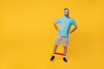Fototapeta na wymiar Full body young smiling happy fitness trainer instructor sporty man sportsman in headband blue t-shirt use fitness elastic bands stand akimbo isolated on plain yellow background Workout sport concept