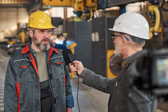 Mature worker in uniform giving an interview to microphone for reporter while they standing in workshop in factory