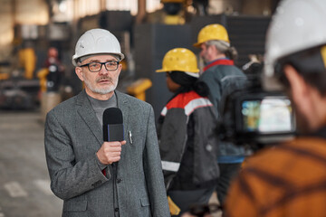 Senior TV journalist in work helmet speaking to microphone during reportage in factory with workers...