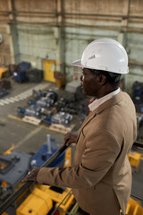 African engineer in work helmet standing on balcony on top floor and watching for the work in...