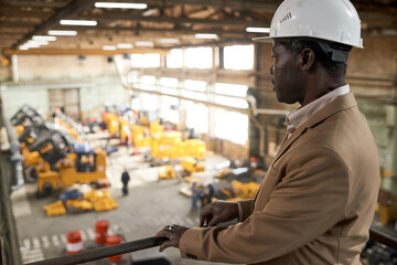 Serious African engineer in work helmet standing on balcony and watching for the work of machines...