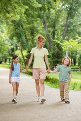 happy woman holding hands with kids while walking in park.