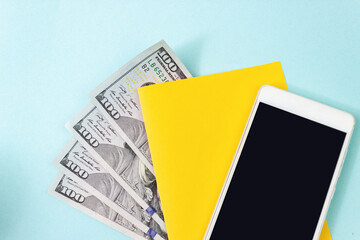 Close up blank yellow piece of paper, 100 dollar bills and empty screen mobile phone on blue background.