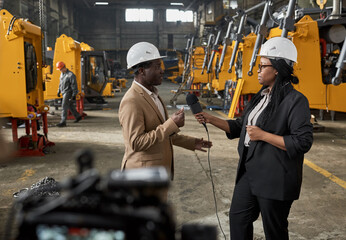 African journalist holding microphone and interviewing engineer in warehouse with new equipment