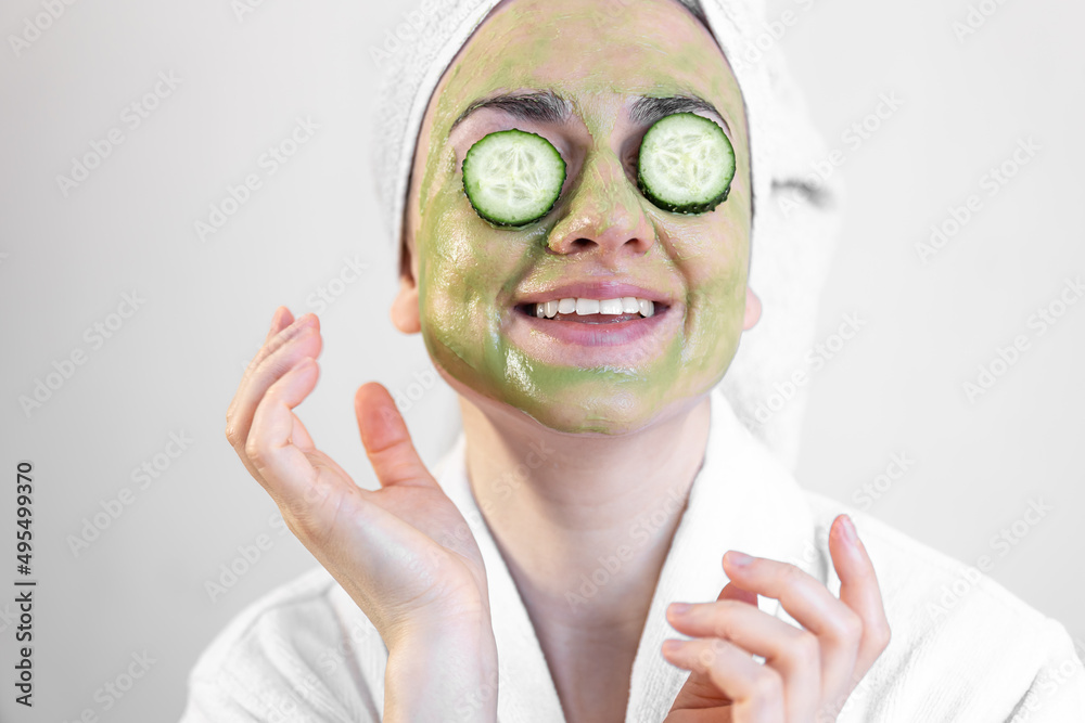 Sticker Young woman with a green face mask and fresh cucumbers in a white bathrobe.