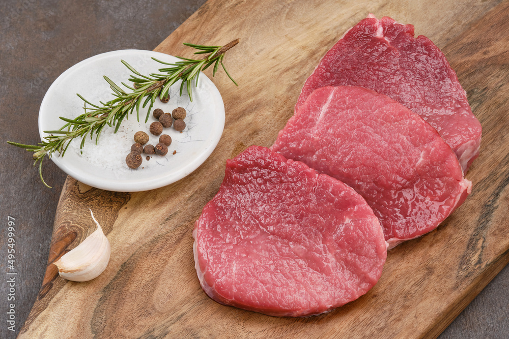 Wall mural Fresh cut of beef on a wooden board with salt, pepper, garlic, rosemary. Raw steak beef meat fillet ready to cook. Selective focus