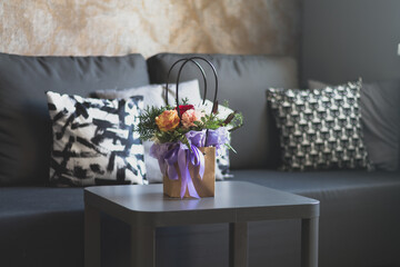 A bouquet of flowers in a paper bag with a purple ribbon stands on a gray table in a room near a dark sofa