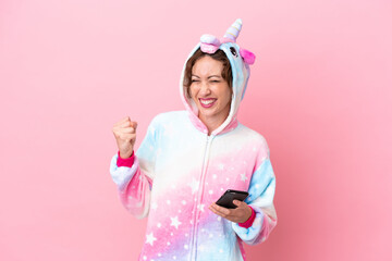 Young caucasian woman with unicorn pajamas isolated on pink background with phone in victory...