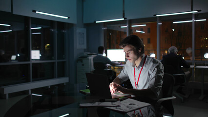 Naklejka na ściany i meble Young businessman type on laptop in dark office with colleagues on background
