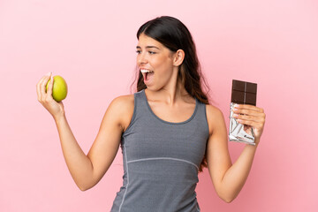 Young caucasian woman isolated on pink background taking a chocolate tablet in one hand and an...