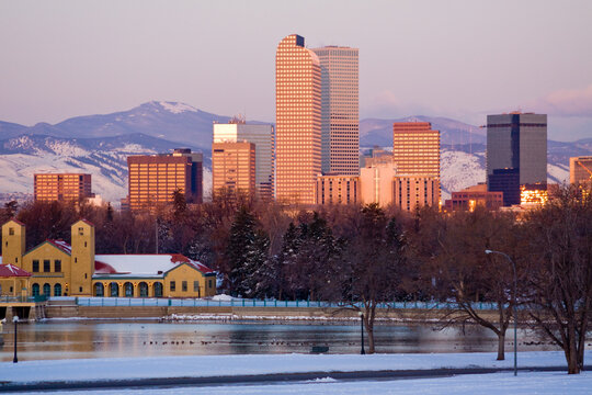 Sunrise turns downtown Denver high rises a golden color as the architecture rises above the trees and lake at Denver city park in winter on this cold Colorado morning.