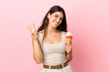 Young caucasian woman with a cornet ice cream isolated on pink background happy and counting three with fingers