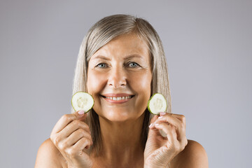Smiling mature woman with two slices of fresh cucumber in hands posing over grey background. Portrait of positive lady with healthy face skin.