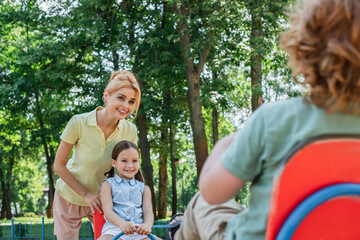 woman smiling near kids riding seesaw on blurred foreground.